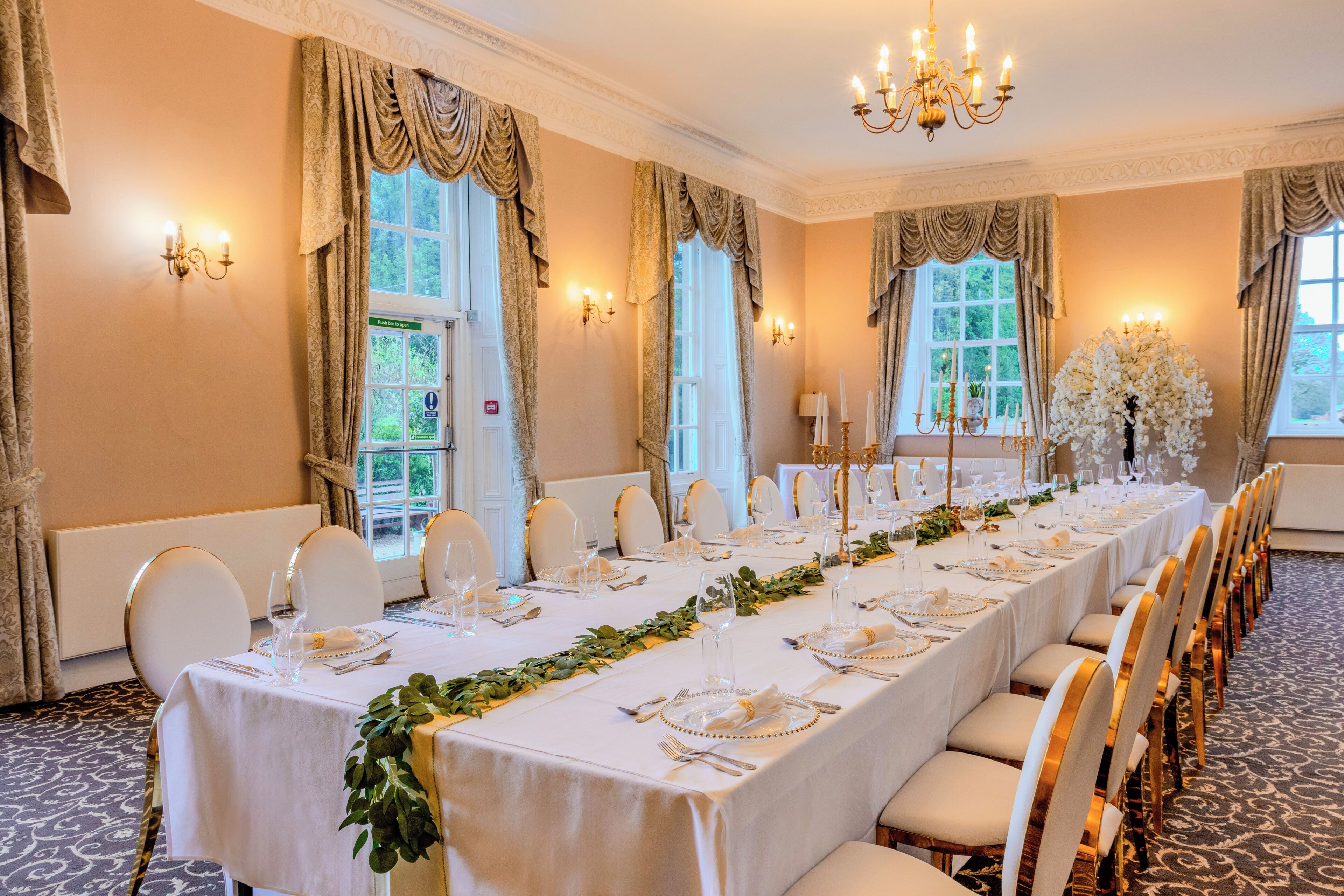 A long banquet table set for a formal event, with white tablecloths, gold accents, and a chandelier.
