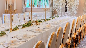Une salle de banquet avec de longues tables dressées pour un événement formel, agrémentée de touches dorées et recouverte de nappes blanches.
