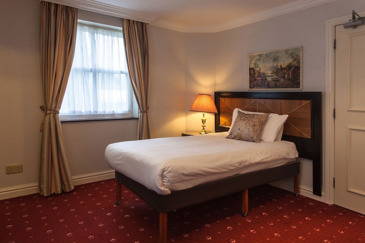 A hotel room with a bed, a nightstand, a lamp, a painting on the wall, and a window with curtains.