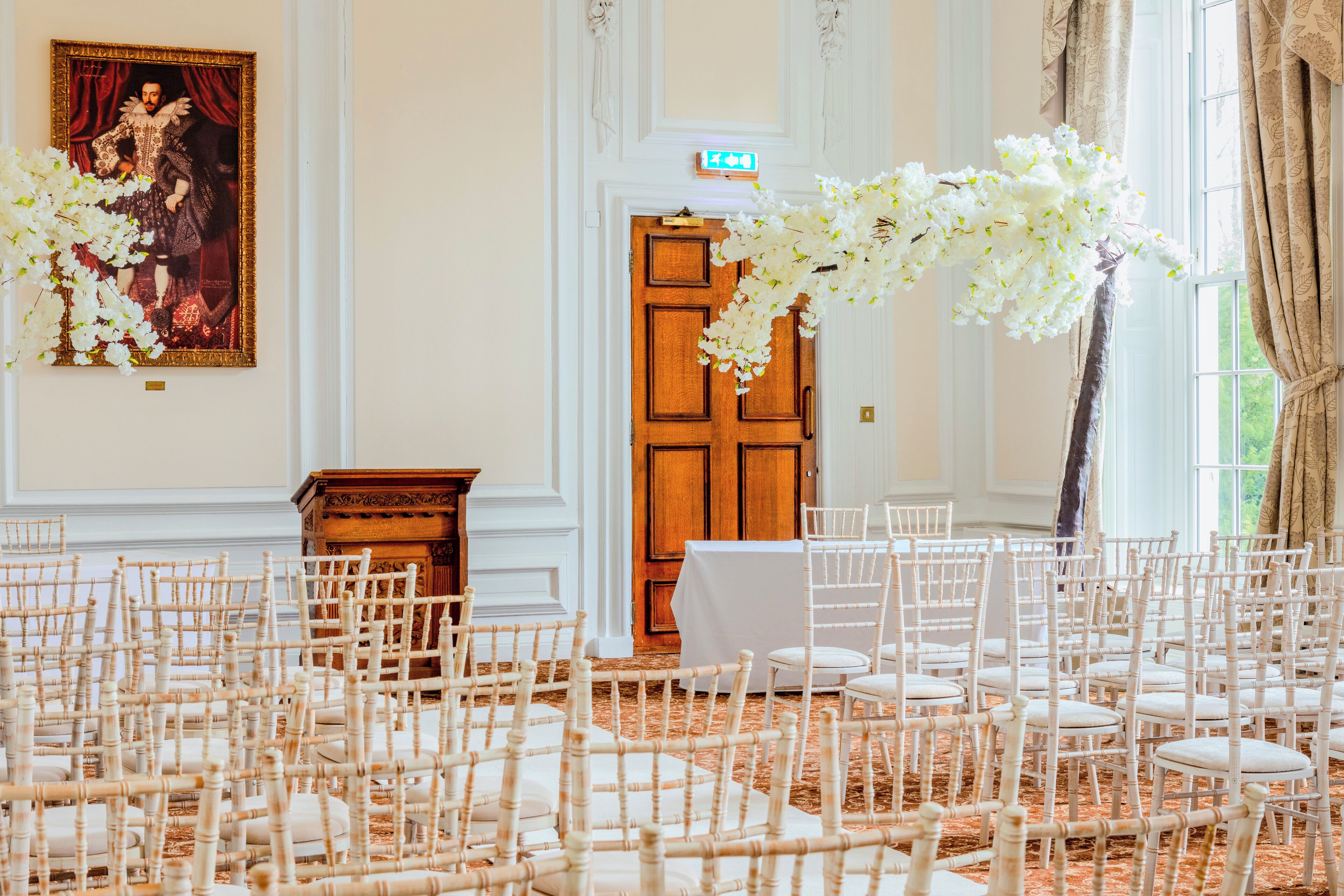 An indoor venue with rows of white chairs, a wooden door, and a large white floral arrangement.