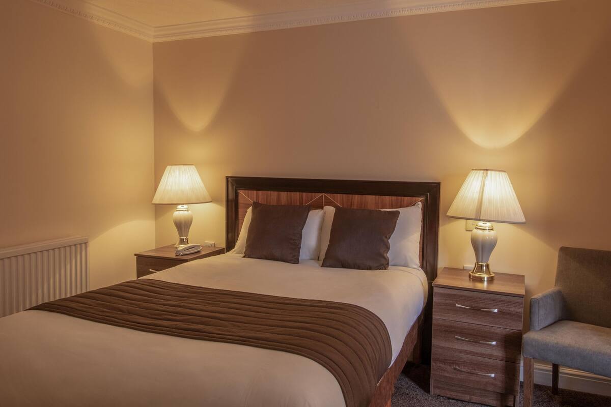 A hotel room with a large bed, two bedside tables with lamps, a chair, and a wooden headboard.