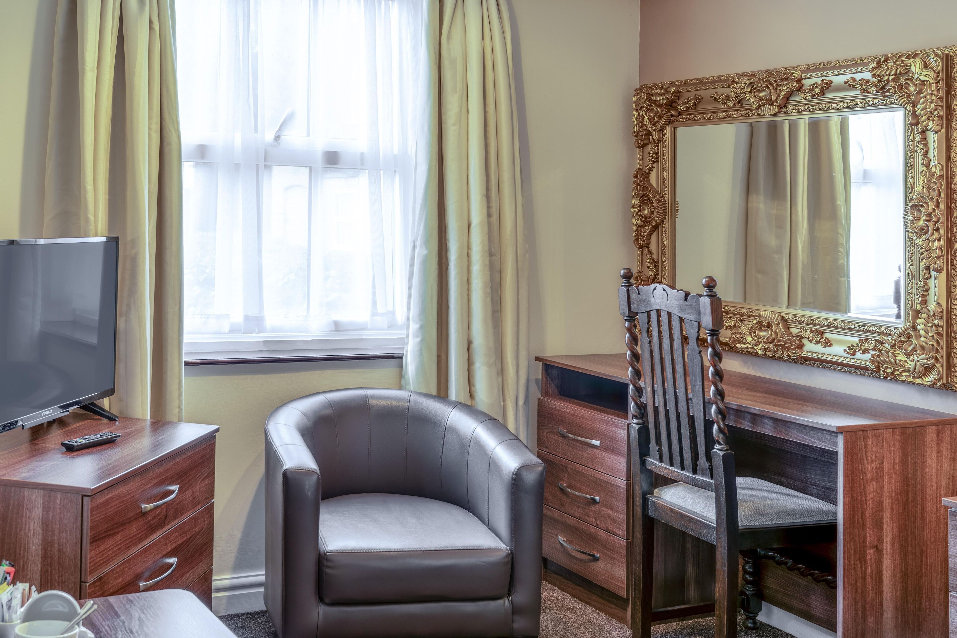 Una habitación de hotel con un sillón de cuero, un aparador de madera con cajones, un televisor y un espejo grande y decorativo.