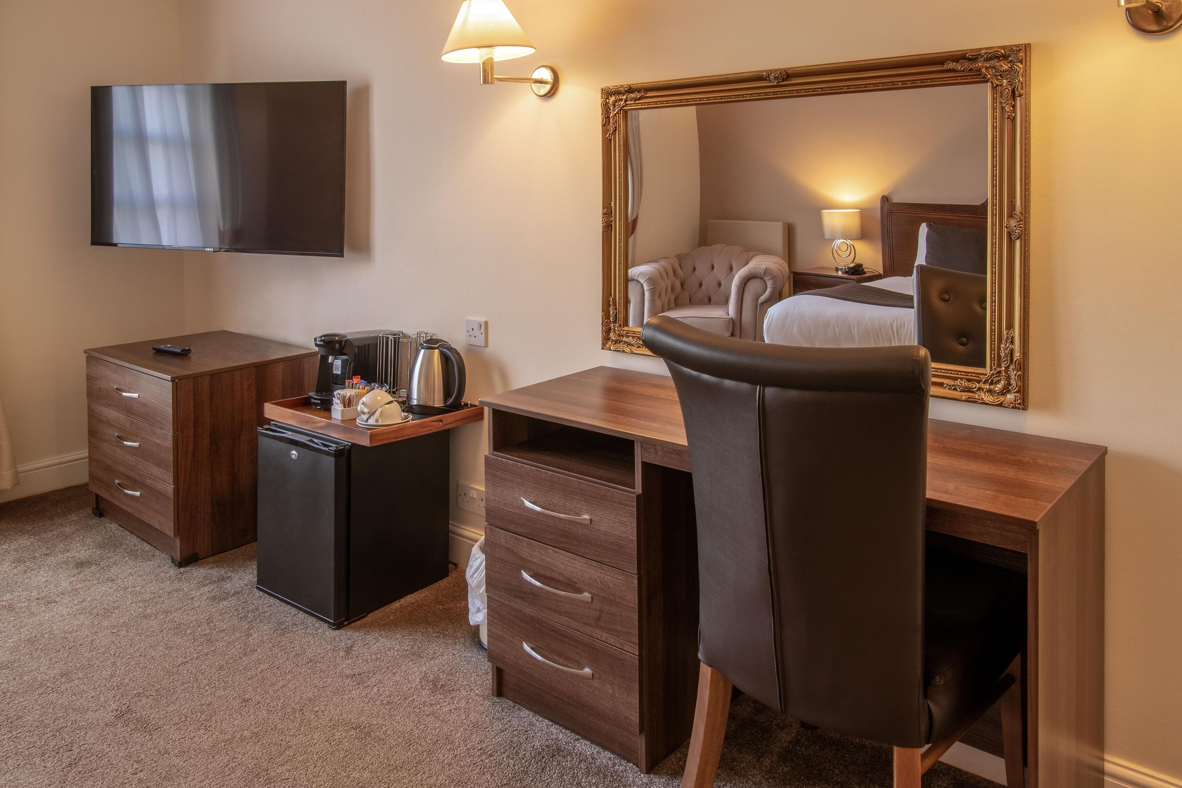 A hotel room with a wooden desk, a black refrigerator, a flat-screen TV, a large mirror, and a comfortable chair.
