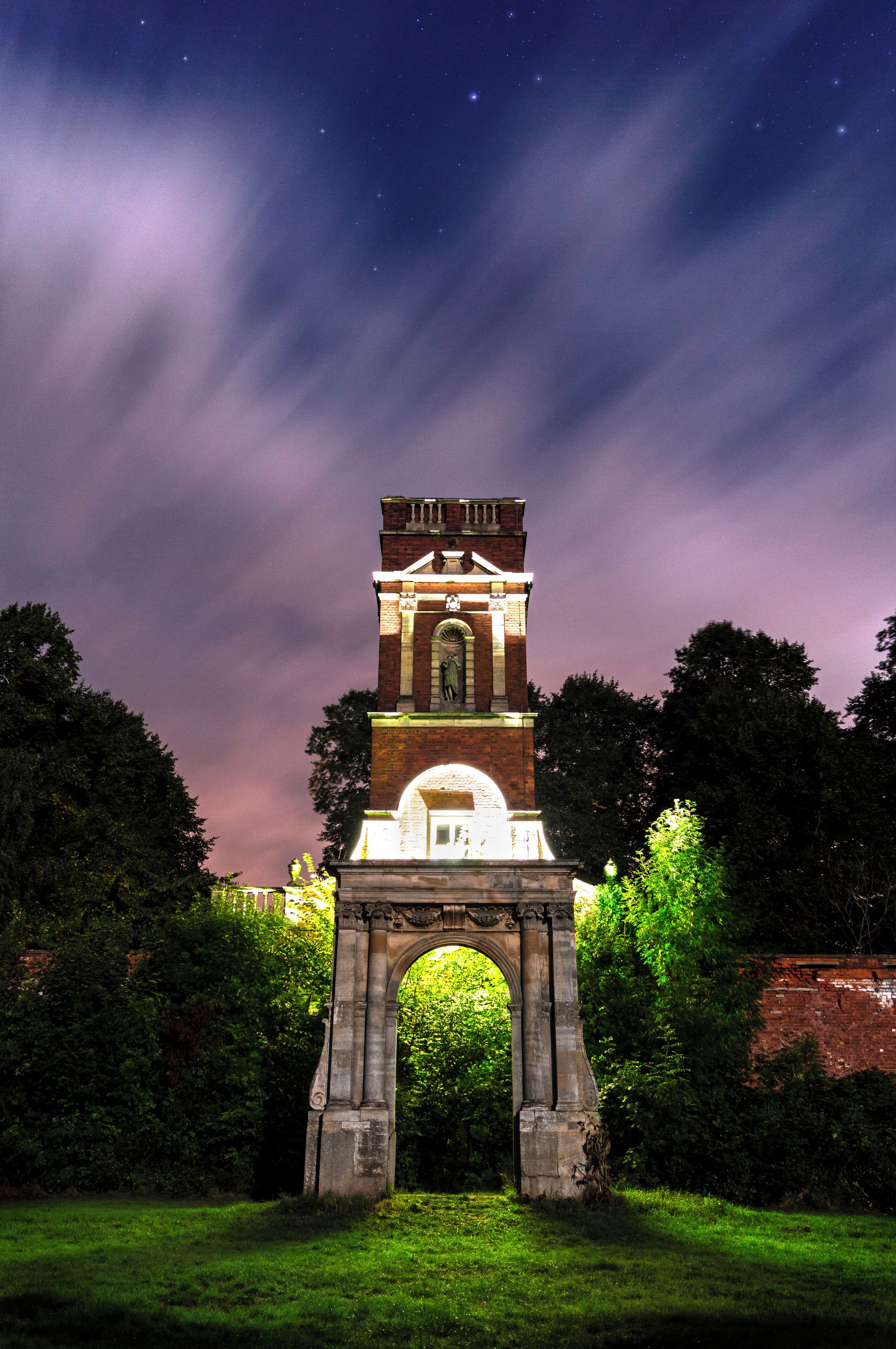 A historic stone archway with a clock tower, surrounded by greenery.