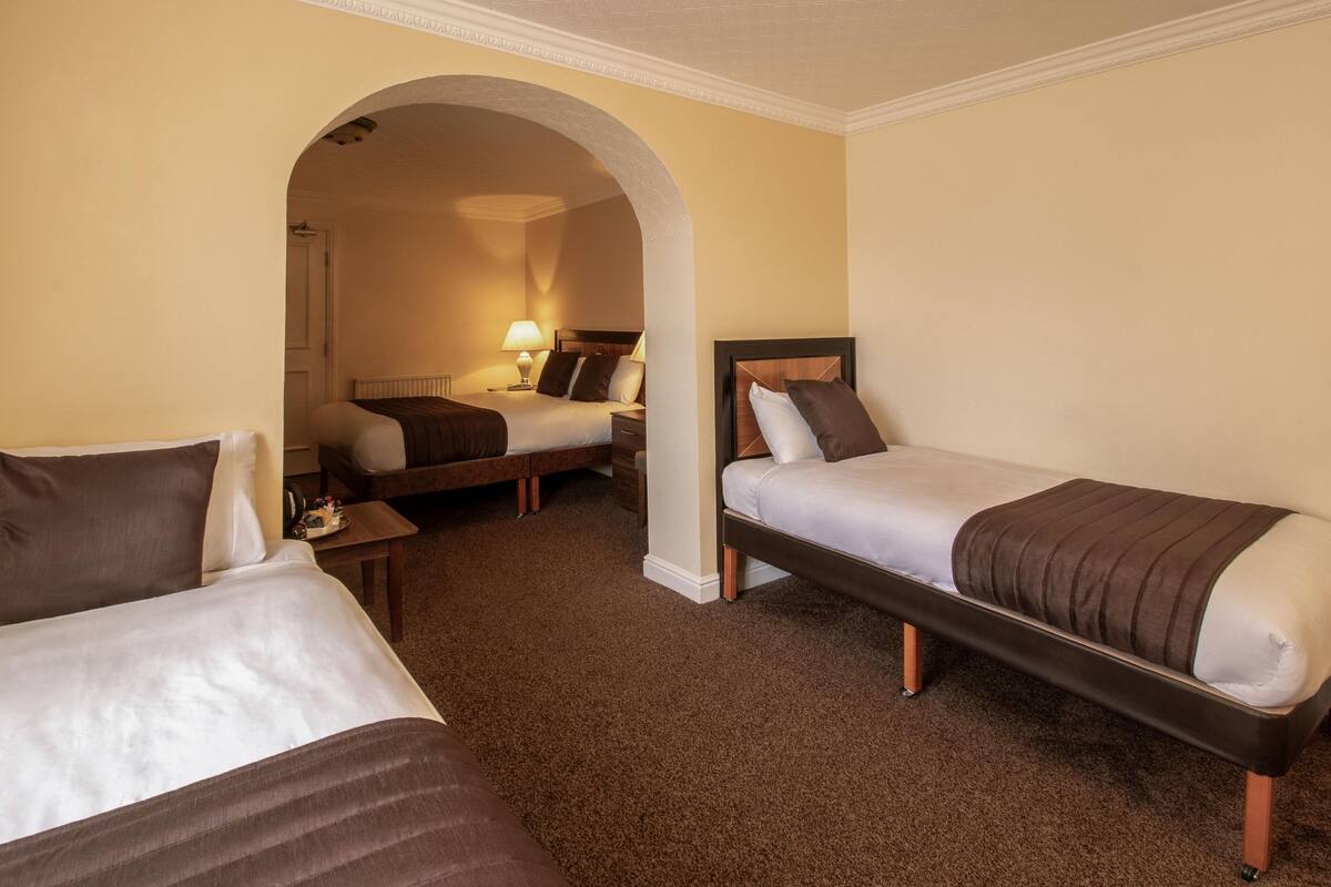 A hotel room with two single beds, a sitting area with a chair and a table, and an arched doorway leading to another room.