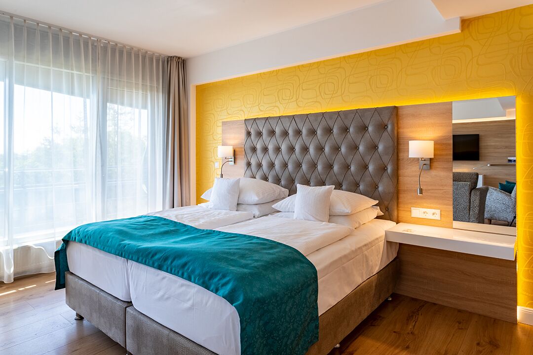 A hotel room with a large bed, a wooden headboard, a bedside table with a lamp, a mirror, and a view of the outside through a window with sheer curtains.