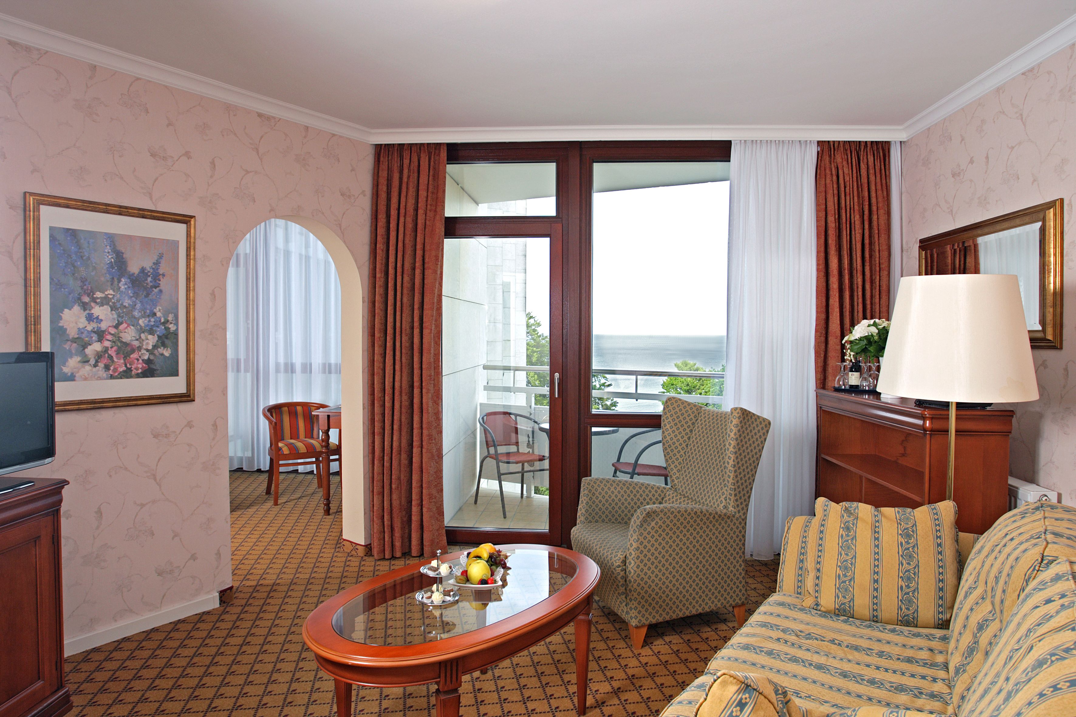 Habitación de hotel con sofá, sillón y una mesa con fruta. Tiene un balcón con una silla y una mesa, además de un televisor sobre un mueble.