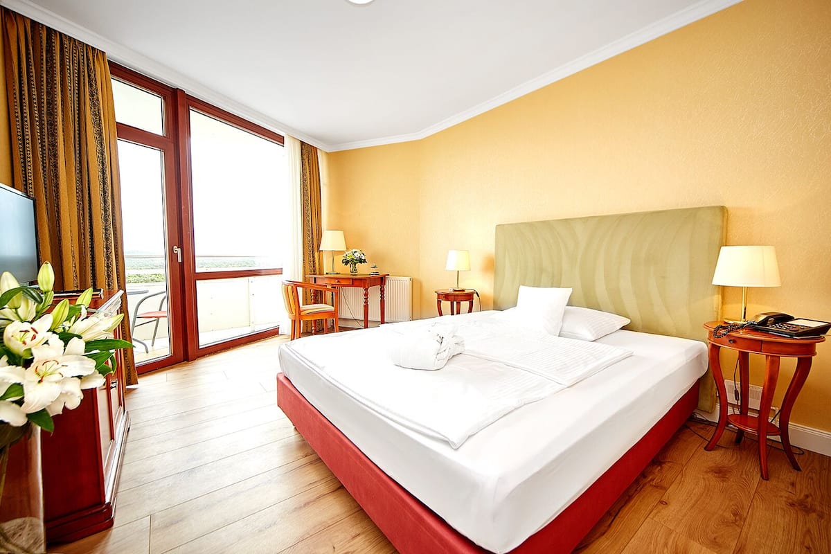 A hotel room with a large bed, a desk, a chair, a small table with a lamp, and a view of the outdoors.