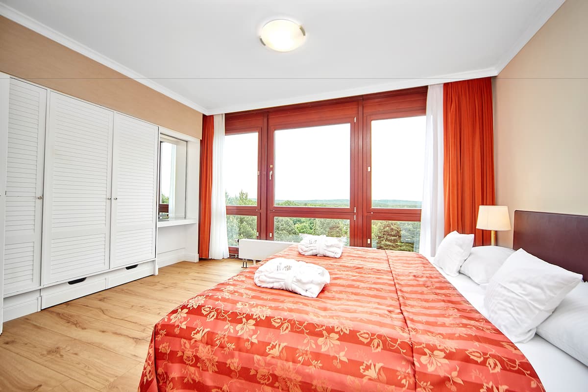A hotel room with a large bed, white curtains, a view of the outdoors, and a white wardrobe.