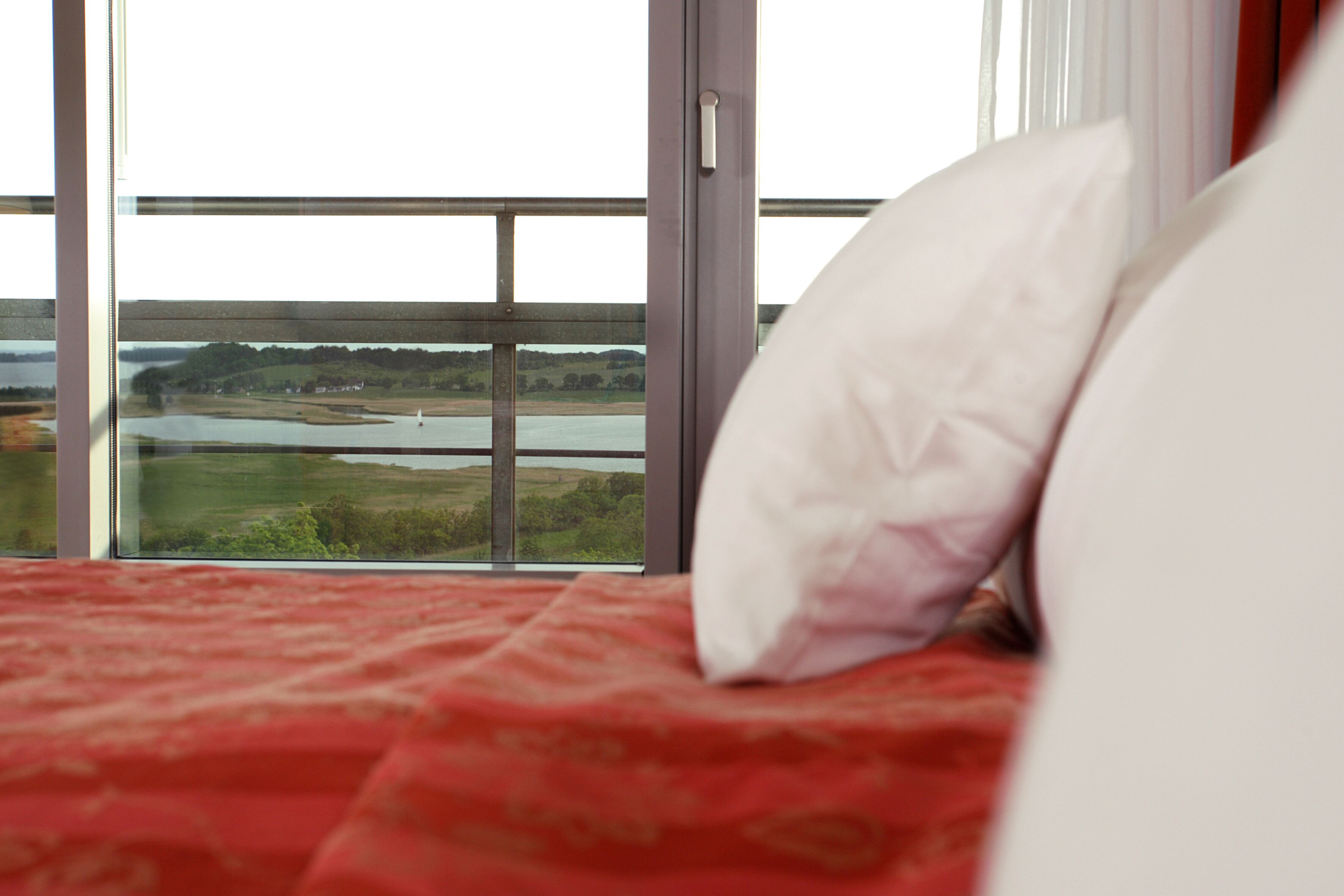 A bed with a red bedspread and a white pillow, a window with a view of a river and landscape, and a door with a handle.