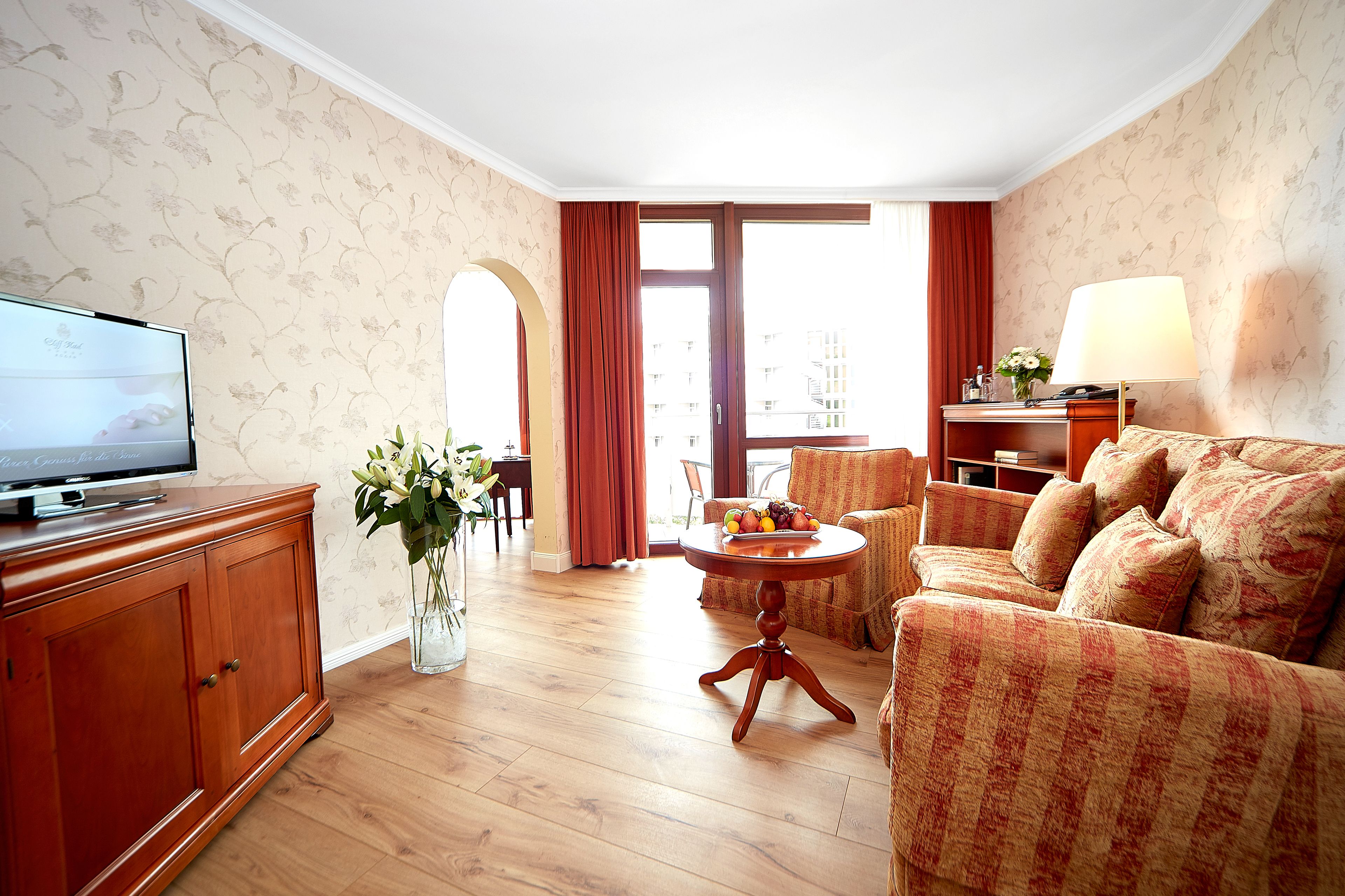 Ein Hotelzimmer mit Holzschrank, Fernseher, rundem Tisch mit Obst, einer Couch und einer Lampe.