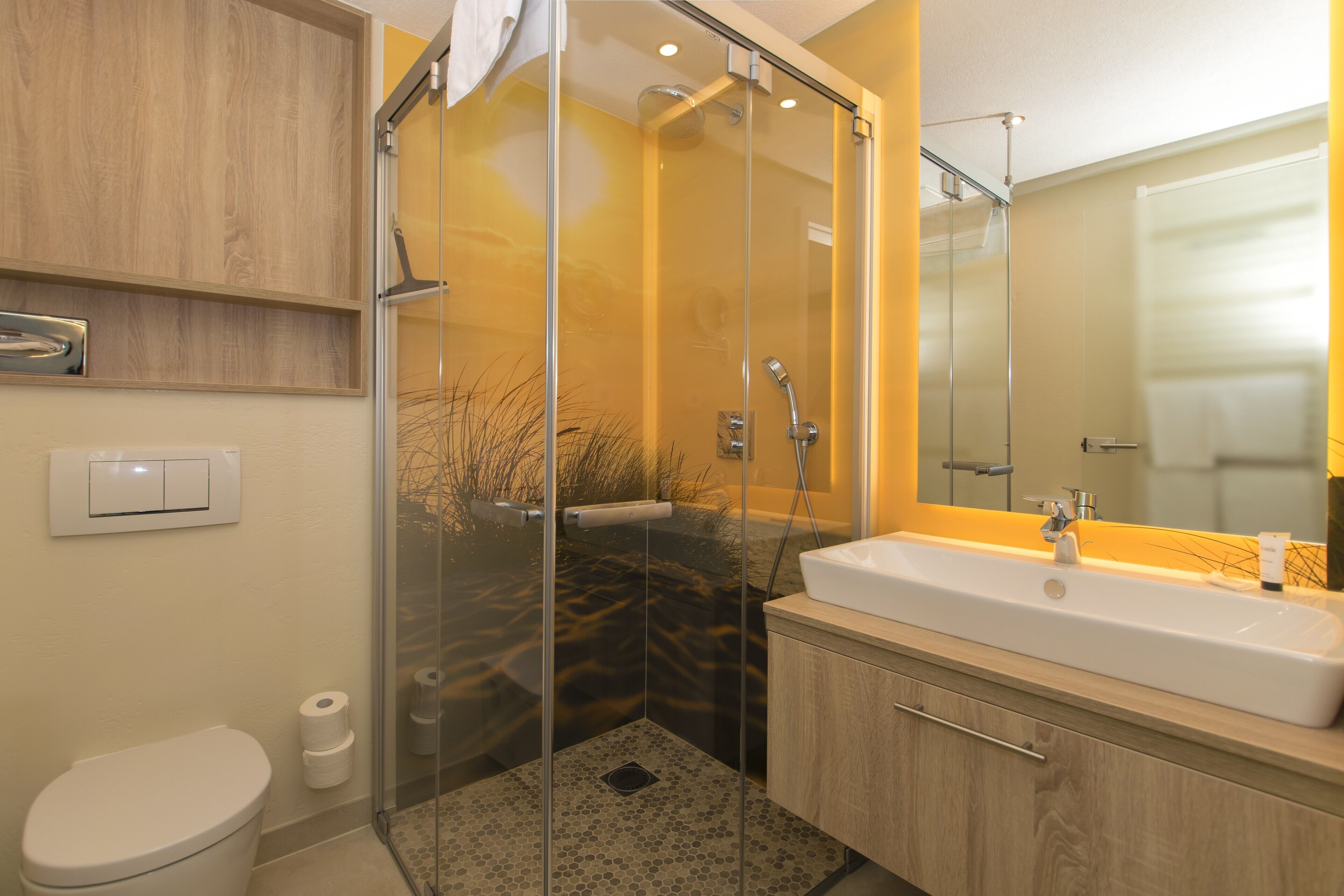 Un baño moderno con ducha envuelta en vidrio, lavamanos blanco y tocador de madera.