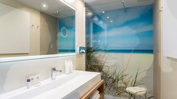 A modern bathroom with a large mirror, a white sink, and a view of the beach through a glass wall.