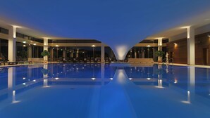 Une grande piscine intérieure dotée d’une architecture moderne, entourée d’espaces aménagés pour se détendre et de plantes en pot.