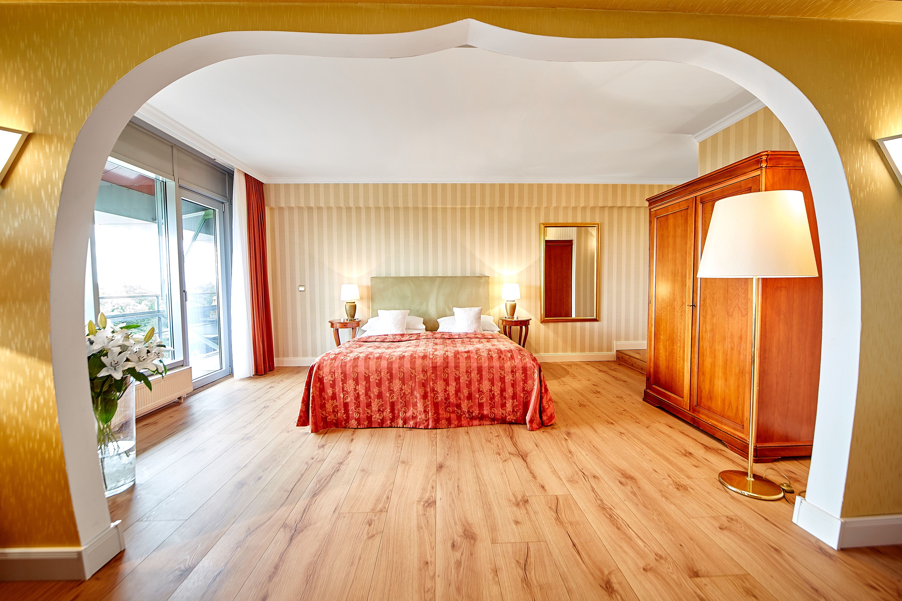 A hotel room with a large bed, a wooden wardrobe, a nightstand with a lamp, and a window with curtains.