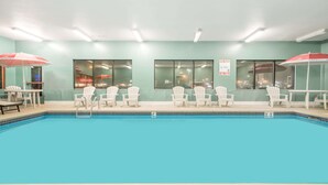 A swimming pool with white chairs and umbrellas, a teal wall, and large windows.