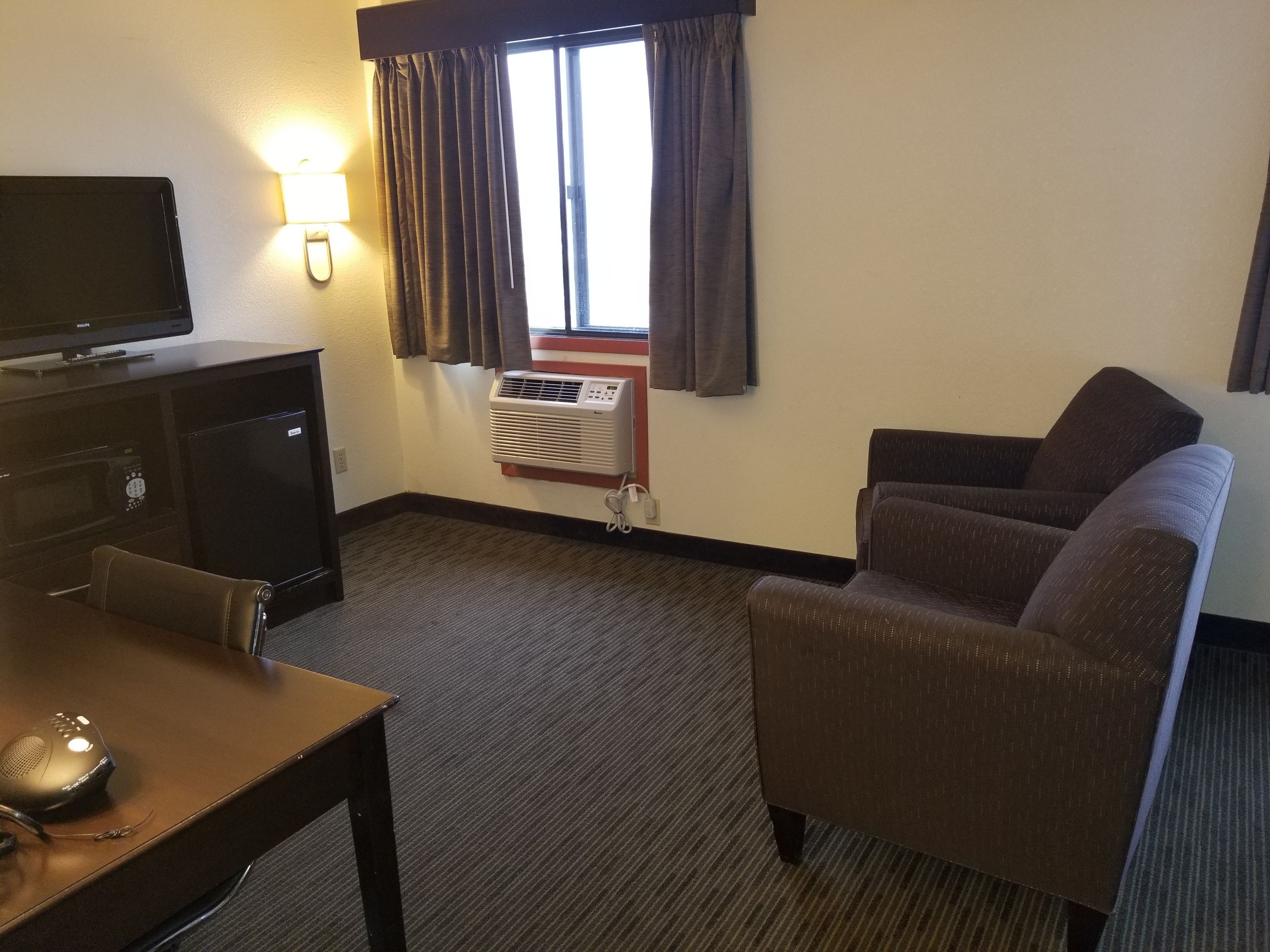 Habitación de hotel con televisión de pantalla plana, escritorio con computadora, sillón marrón, sofá y ventana con cortinas.