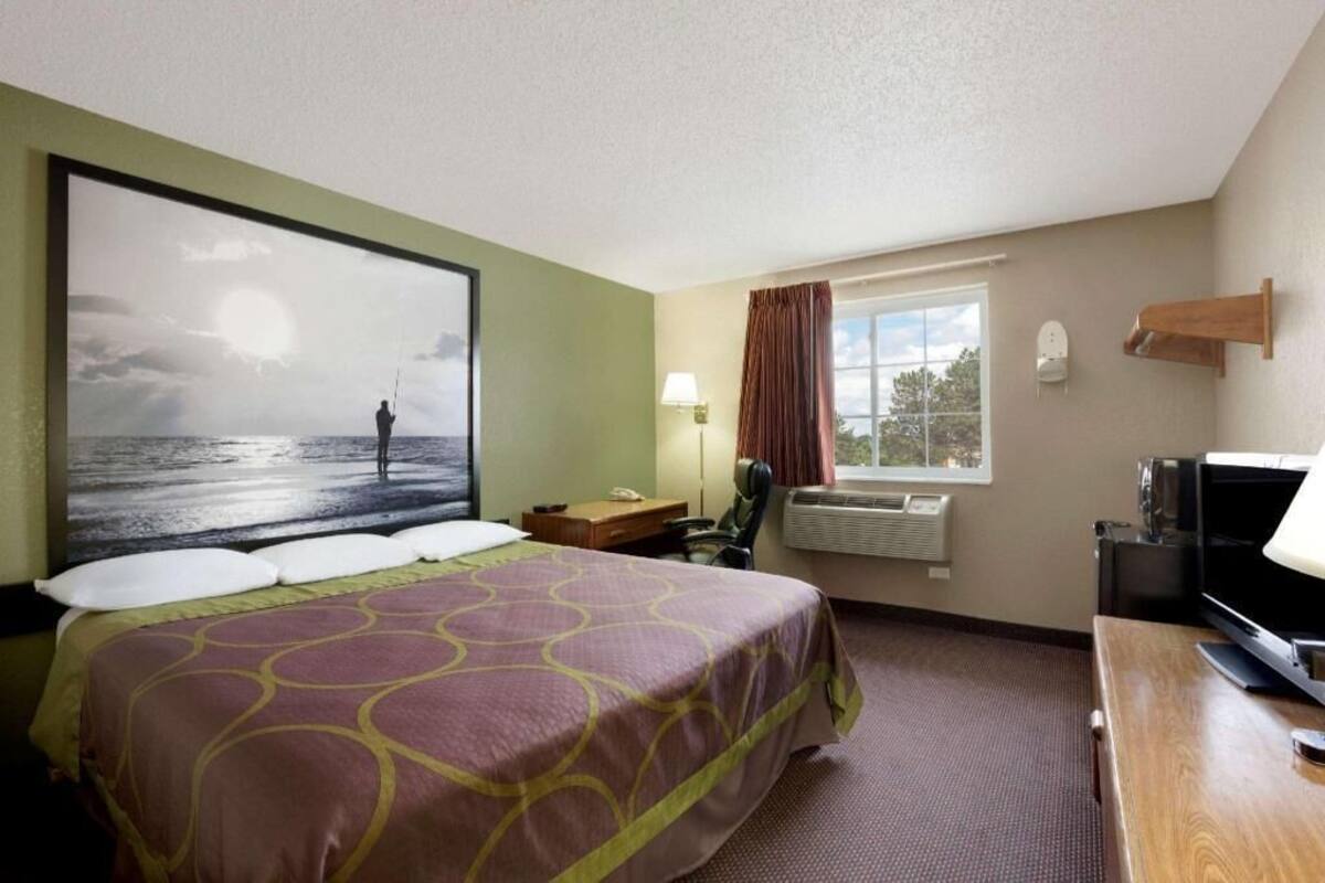 Habitación de hotel con cama, escritorio, televisor, ventana con cortinas, calefactor en la pared y un cuadro grande de una persona pescando.