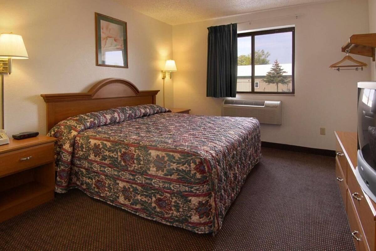 Habitación de hotel con cama, lámparas de noche, televisión, aire acondicionado y ventana con cortinas.