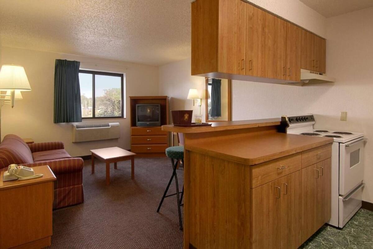 Habitación de hotel compacta con kitchenette, sofá, una mesita, televisor y ventana con cortinas.