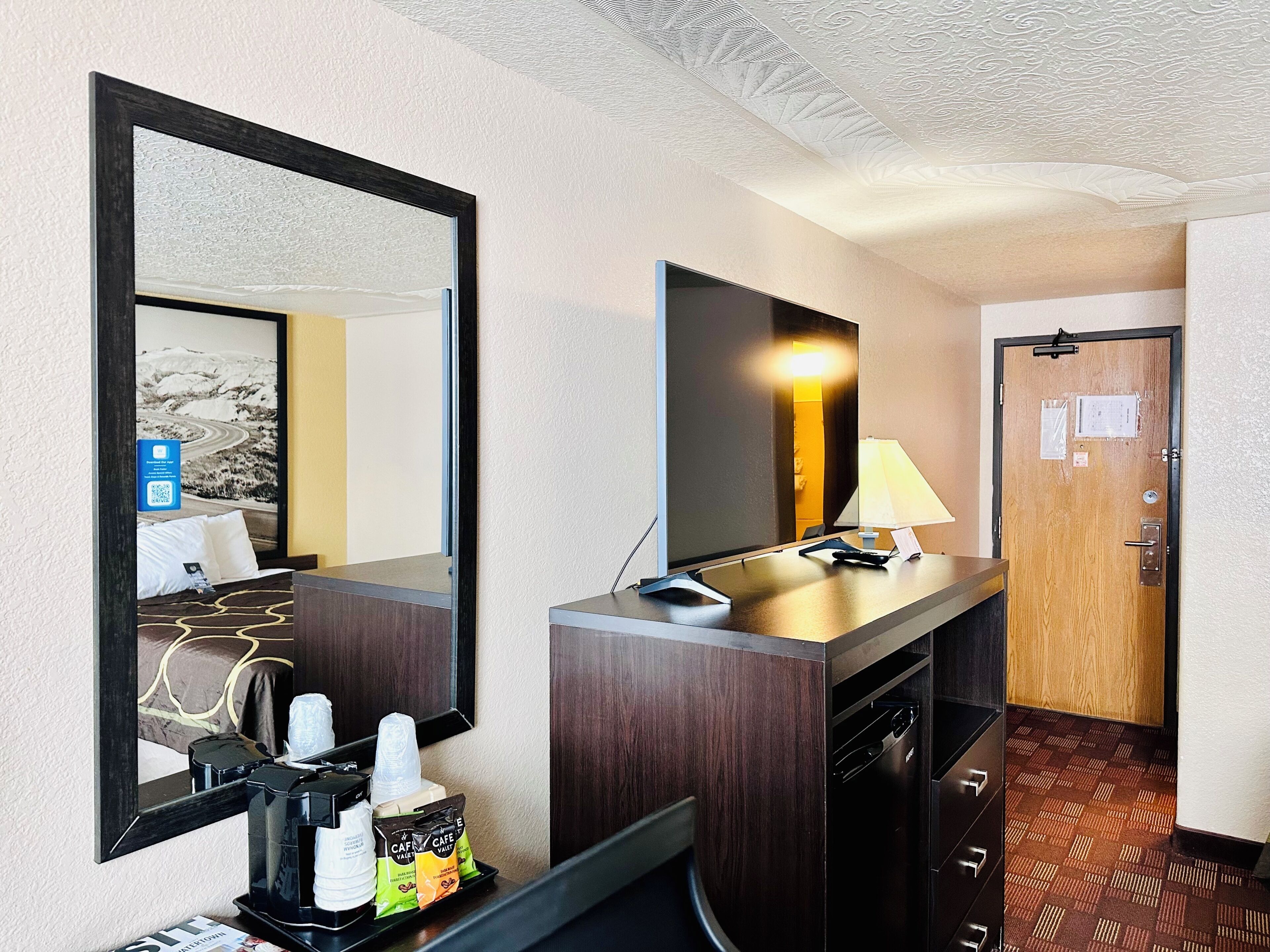 Une chambre d’hôtel dotée d’un grand miroir, d’un téléviseur à écran plat, d’un bureau avec une lampe, d’une machine à café et d’un mini-bar.