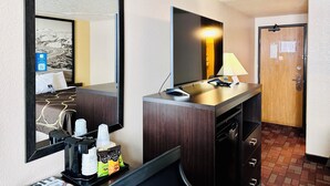 Habitación de hotel con un espejo grande, un televisor de pantalla plana, un escritorio con lámpara, cafetera y minibar.