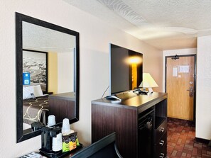 Ein Hotelzimmer mit einem großen Spiegel, einem Flachbildfernseher, einem Schreibtisch mit Lampe, einer Kaffeemaschine und einer Minibar.