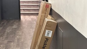 Dos cajas de cartón con la etiqueta 'Pickleball Net' apoyadas contra una pared en un pasillo con suelo de madera.