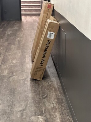 Zwei mit 'Pickleball Netz' beschriftete Kartons lehnen an einer Wand in einem Korridor mit Holzboden.