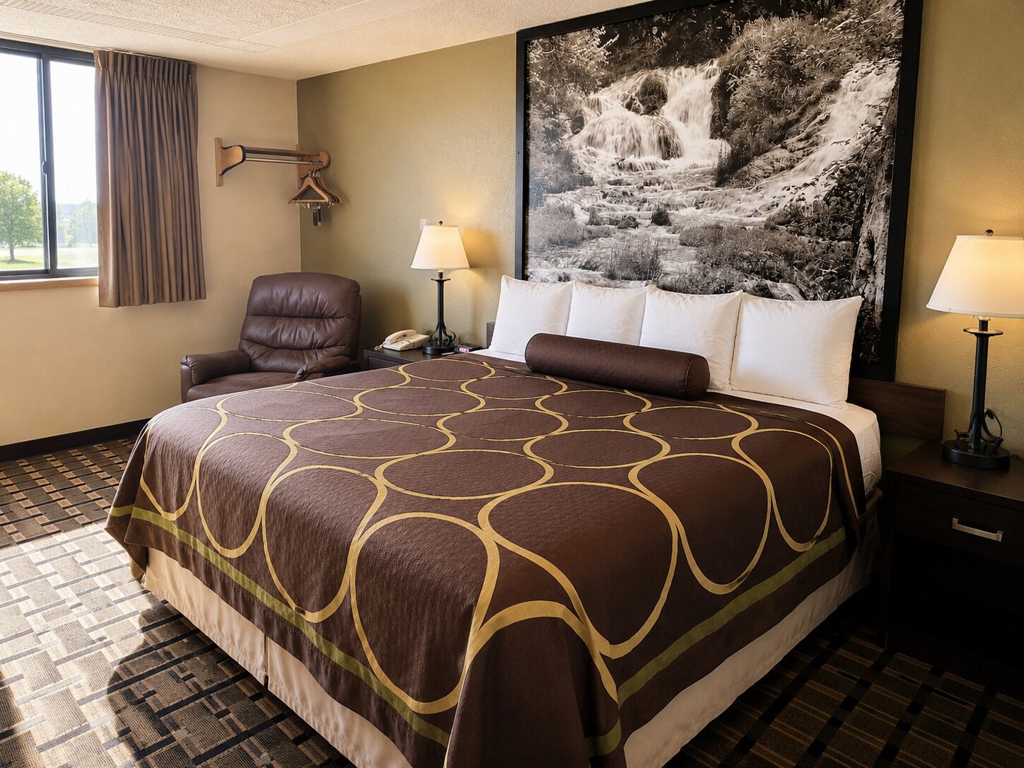 Une chambre d’hôtel avec un grand lit, une chaise, deux tables de chevet avec des lampes, un grand tableau encadré au mur et une fenêtre avec des rideaux.