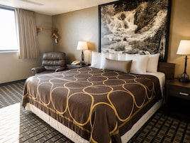 A hotel room with a large bed, a chair, a desk, a lamp, a telephone, and a painting of a waterfall.