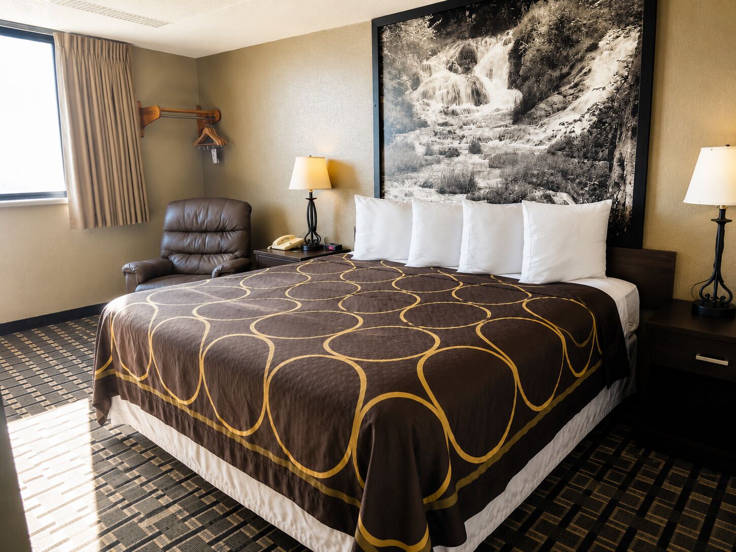 A hotel room with a large bed, a chair, a desk with a lamp, a telephone, and a painting of a waterfall on the wall.