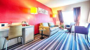 A hotel room with a red accent wall, a sofa, a chair, a small table with a lamp, and a window with curtains. - Leonardo Hotel Mannheim - Ladenburg (Ladenburg)