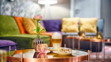 Ein runder Couchtisch mit Glasplatte, eine Vase mit einer rosa Blume, ein Glas bernsteinfarbener Flüssigkeit und ein Teller mit einem Gebäckstück.
