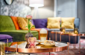 A round coffee table with a glass top, a pink flower vase, a glass of amber liquid, and a plate with a pastry. - Leonardo Hotel Mannheim - Ladenburg (Ladenburg)