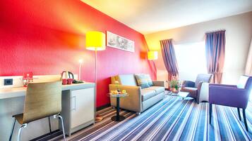A hotel room with a striped carpet, a sofa, a chair, a desk with a lamp, and a window with curtains.
