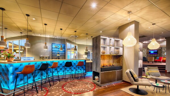 A bar area with a blue cushioned bar counter, multiple bar stools, and a fireplace. - Leonardo Hotel Mannheim - Ladenburg (Ladenburg)