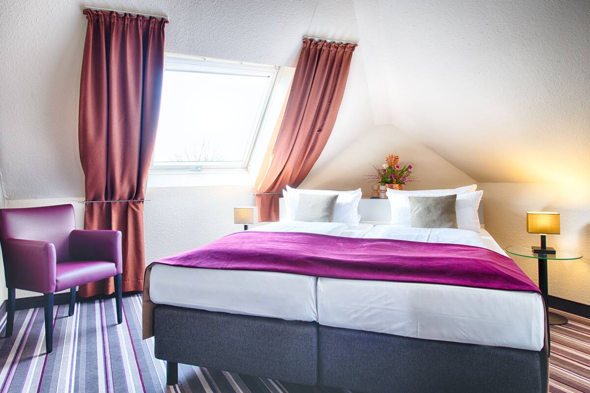 A hotel room with a large bed, a chair, a nightstand, and a window with curtains.