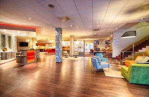 A spacious lobby with a wooden floor, a reception desk, seating area with chairs, and a staircase leading to upper floors.
