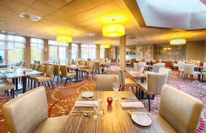 A spacious dining area with multiple tables and chairs, large windows allowing natural light, and a patterned carpet. - Leonardo Hotel Mannheim - Ladenburg (Ladenburg)