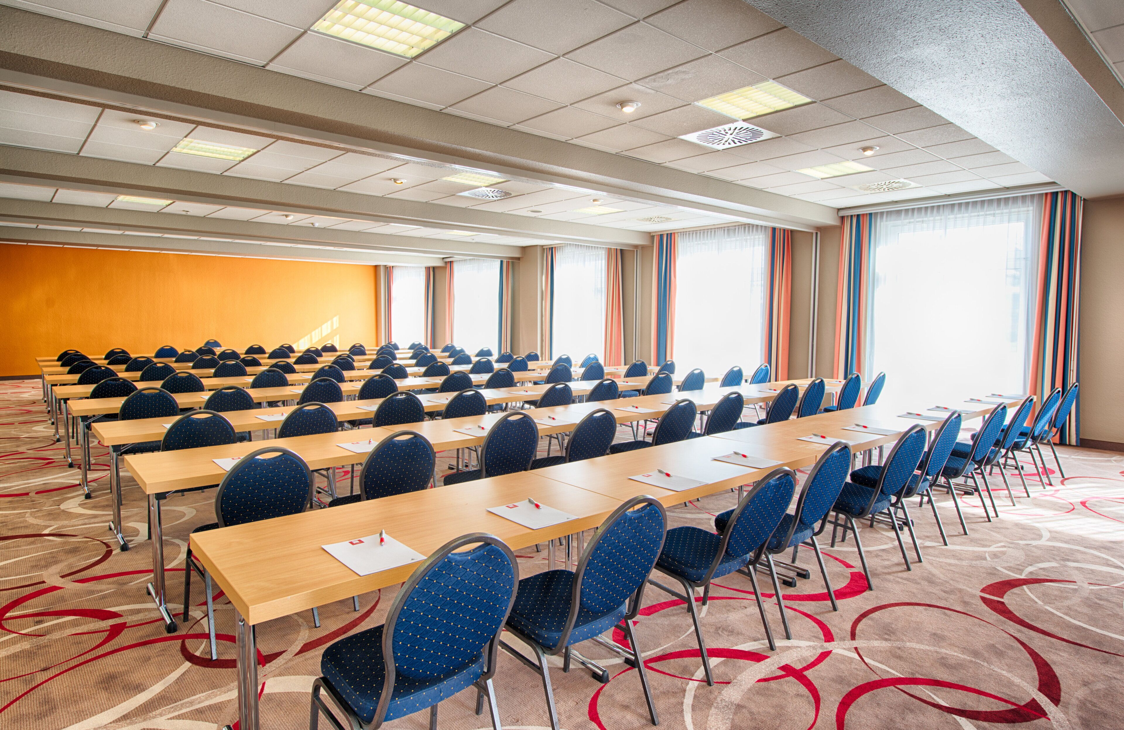 Un salón de conferencias con mesas y sillas en filas, amplias ventanas con cortinas y piso alfombrado.