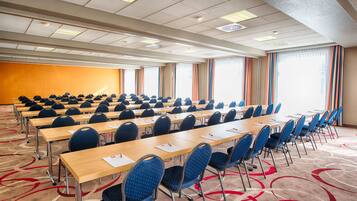Un salón de conferencias con mesas y sillas en filas, amplias ventanas con cortinas y piso alfombrado.