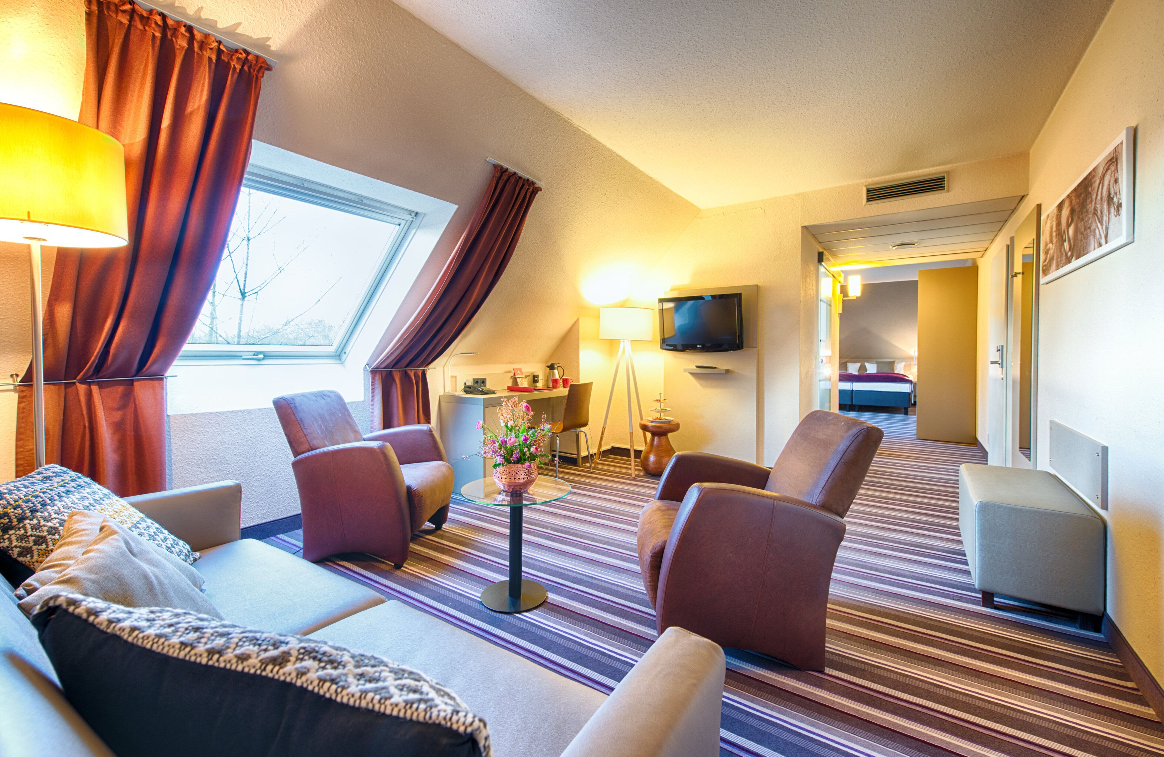 A hotel room with a sofa, armchairs, a TV, a small table with a vase, and a window with curtains.