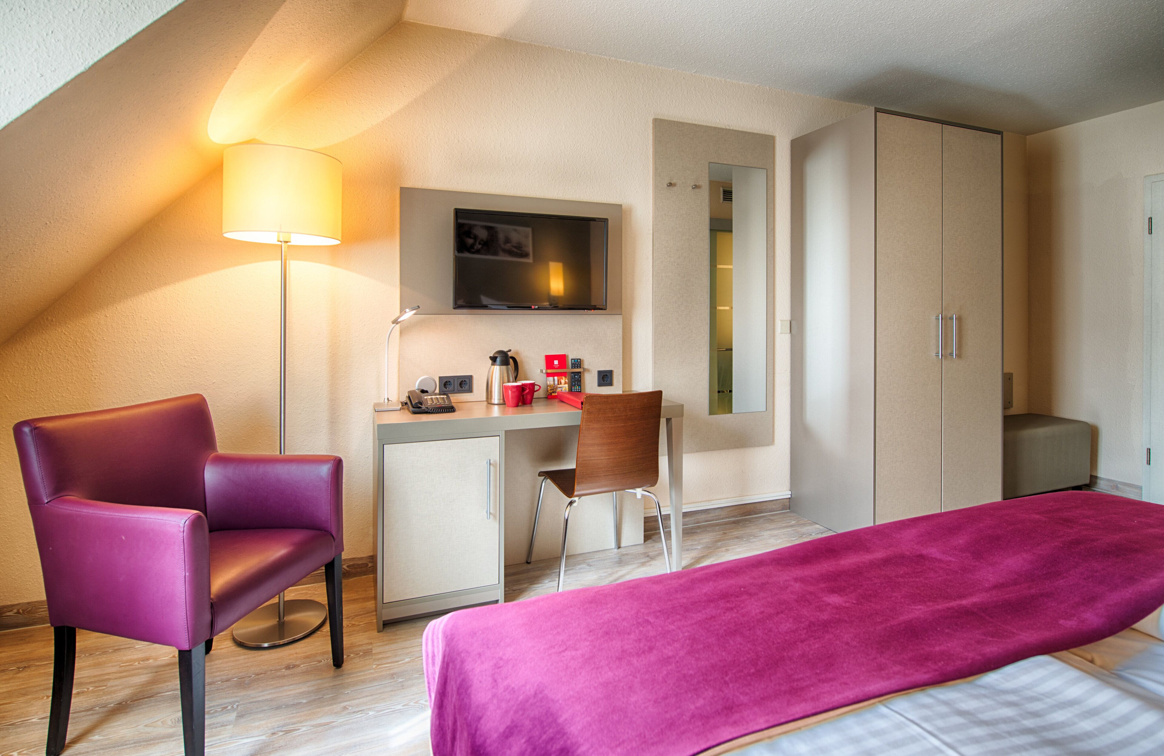 A hotel room with a bed, a desk with a chair, a television, and a wardrobe.