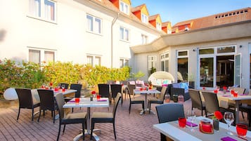 Un espacio exterior para comer con mesas y sillas, una estatua de Buda, y un edificio con varias ventanas y balcones.
