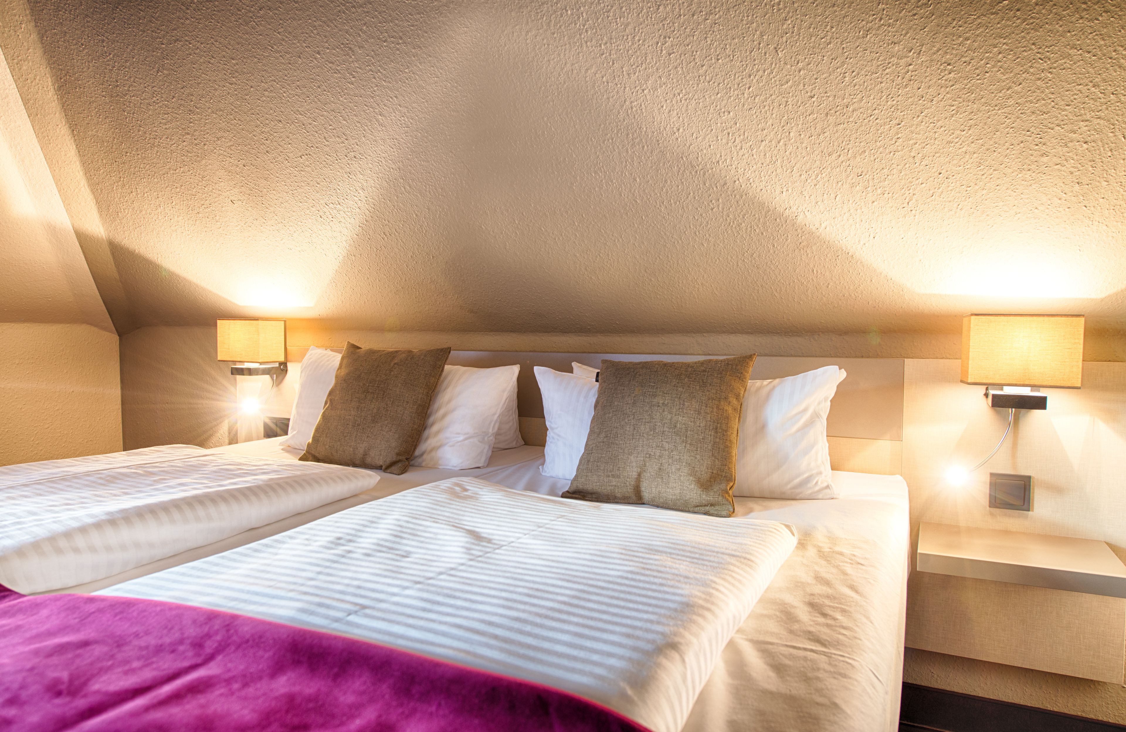Habitación de hotel con dos camas, una colcha rosa y dos lámparas adosadas a la pared.