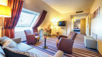 A hotel room with a sofa, armchairs, a TV, a small table with a vase, and a window with curtains.