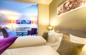 A hotel room with a large bed, a sofa, a desk, and two chairs. There are two lamps on the wall and a painting above the bed. - Leonardo Hotel Mannheim - Ladenburg (Ladenburg)
