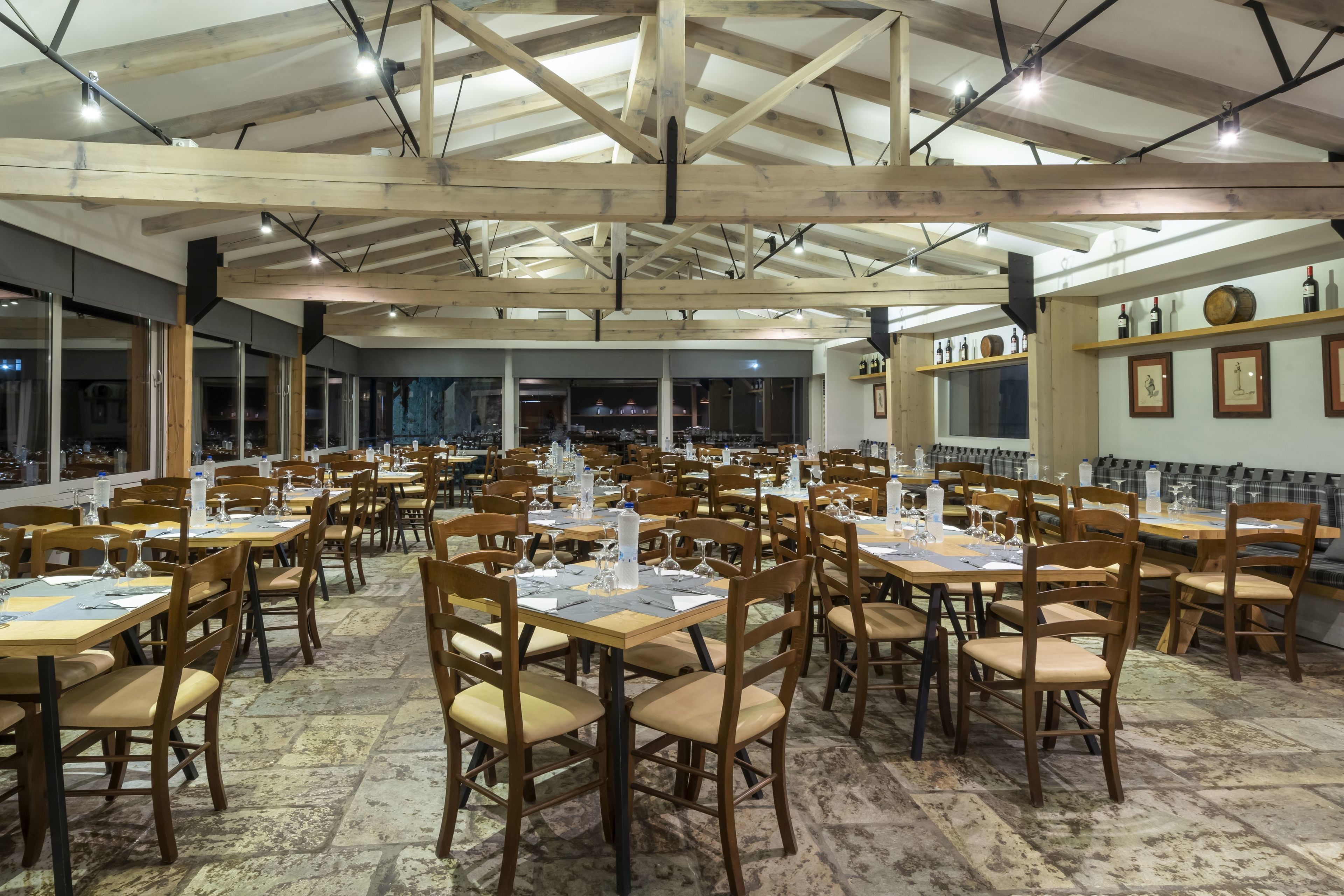 A spacious restaurant with wooden tables and chairs, large windows, and a high ceiling with exposed beams.
