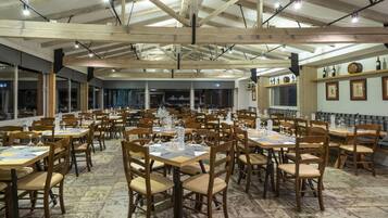 A spacious restaurant with wooden tables and chairs, large windows, and a high ceiling with exposed beams.